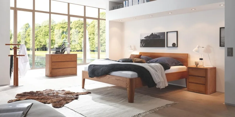 Helles Schlafzimmer mit Holzbett, gemusterter Bettwäsche und zwei Nachttischen vor einer Fensterfront.