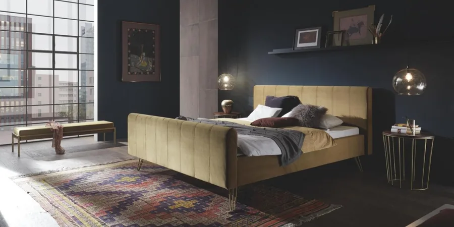 Schlafzimmer mit senfgelbem Polsterbett, gemusterter Bettwäsche und Beistelltisch vor dunkler Wand.