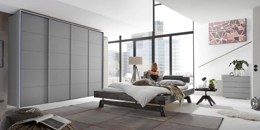 Schlafzimmer mit grau gepolstertem Bett, Kleiderständer und Kommode vor einer Fensterfront mit Seeblick.