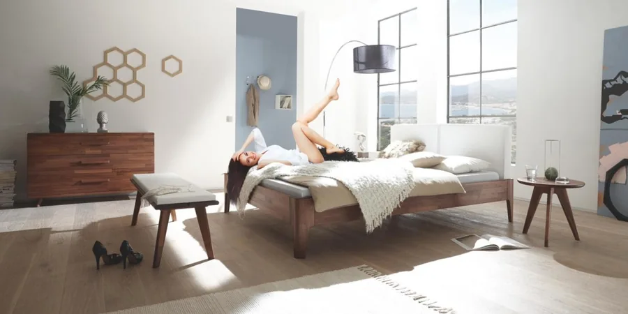 Helles Schlafzimmer mit Holzbett, Sitzhocker und Kommode vor großen Fenstern.