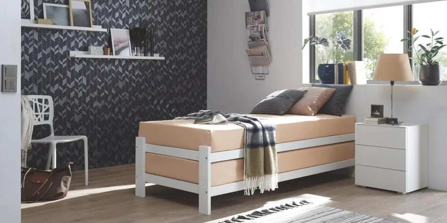 Jugendzimmer mit weißem Bett, weißer Kommode und dunkel gemusterter Tapete.