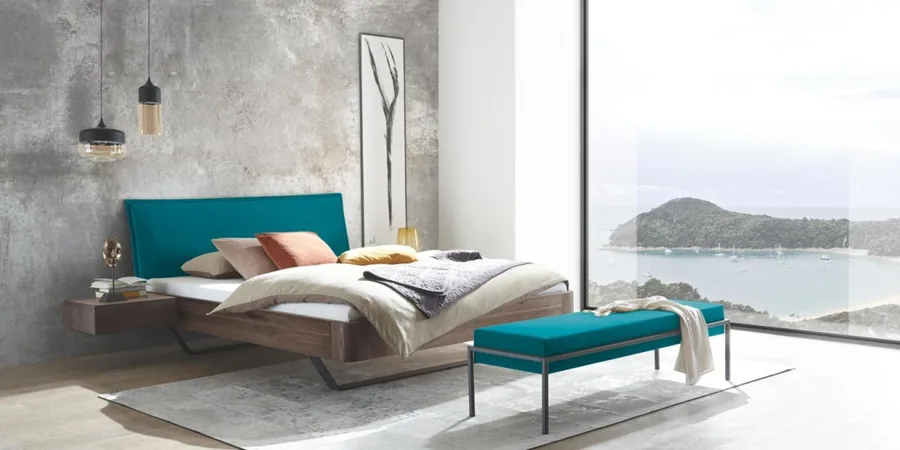 Schlafzimmer mit Holzbett, türkisem Kopfteil und türkiser Bank vor einer Fensterfront mit Meerblick.