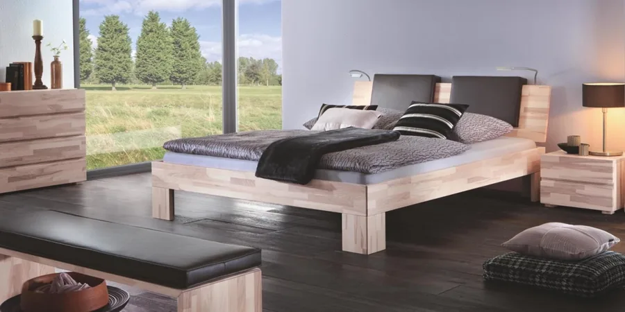 Massives Holzbett mit dunkler Decke, Lederbank am Fußende und Nachttisch vor heller Wand.