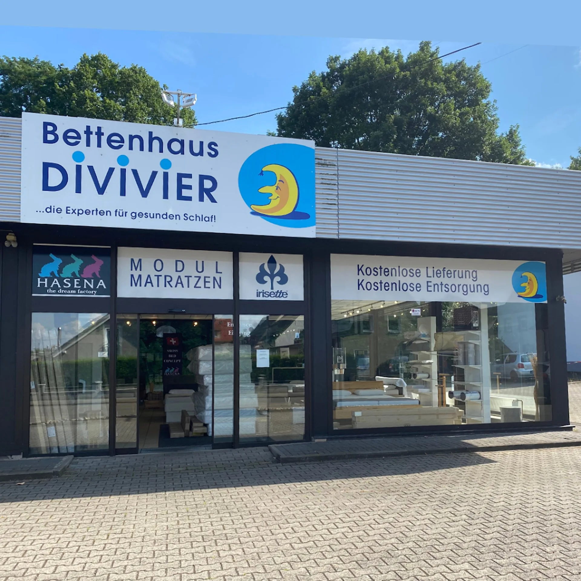 Das Ladengeschäft von Bettenhaus Divivier: eine Glasfront mit blauem Schriftzug und dem Mond-Logo, daneben Schilder für Hasena, Modul-Matratzen, kostenlose Lieferung und kostenlose Entsorgung.